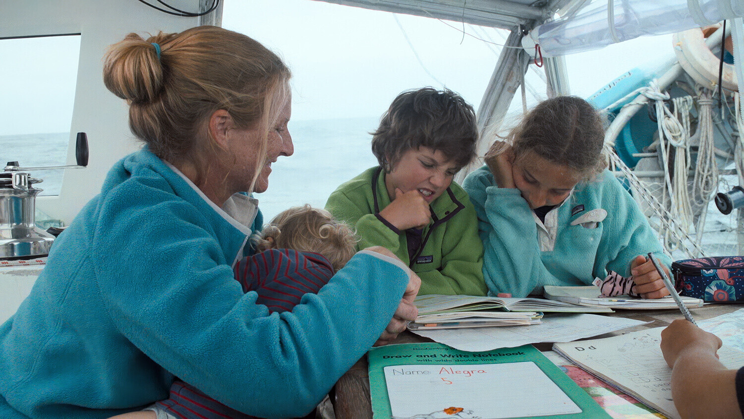 Sabine Schw&ouml;rer homeschooling three children on the deck of the TOPtoTOP expedition sailboat while holding a baby &ndash; school workbooks spread on the table