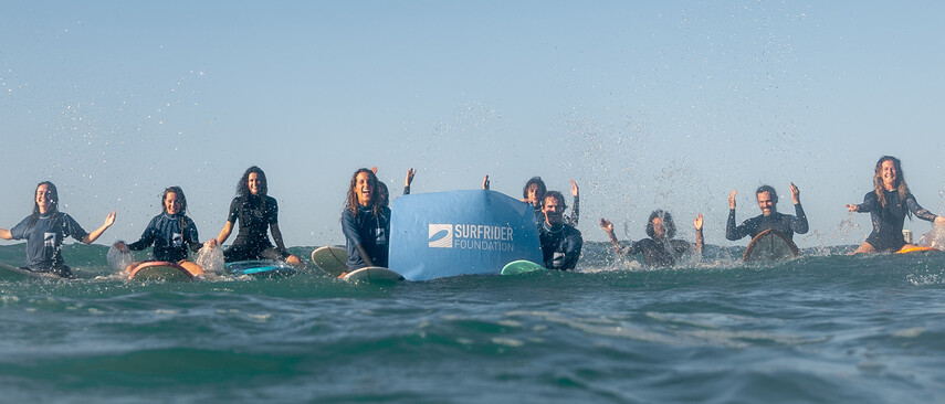 A group sitting on surfboards holding up a blue banner with the Surfrider Foundation logo